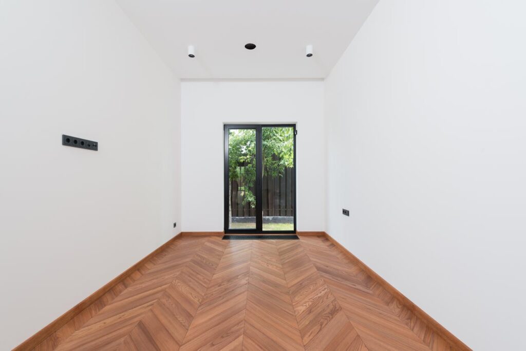 A clean, minimalist interior room with white walls, wooden parquet flooring, and a glass door.